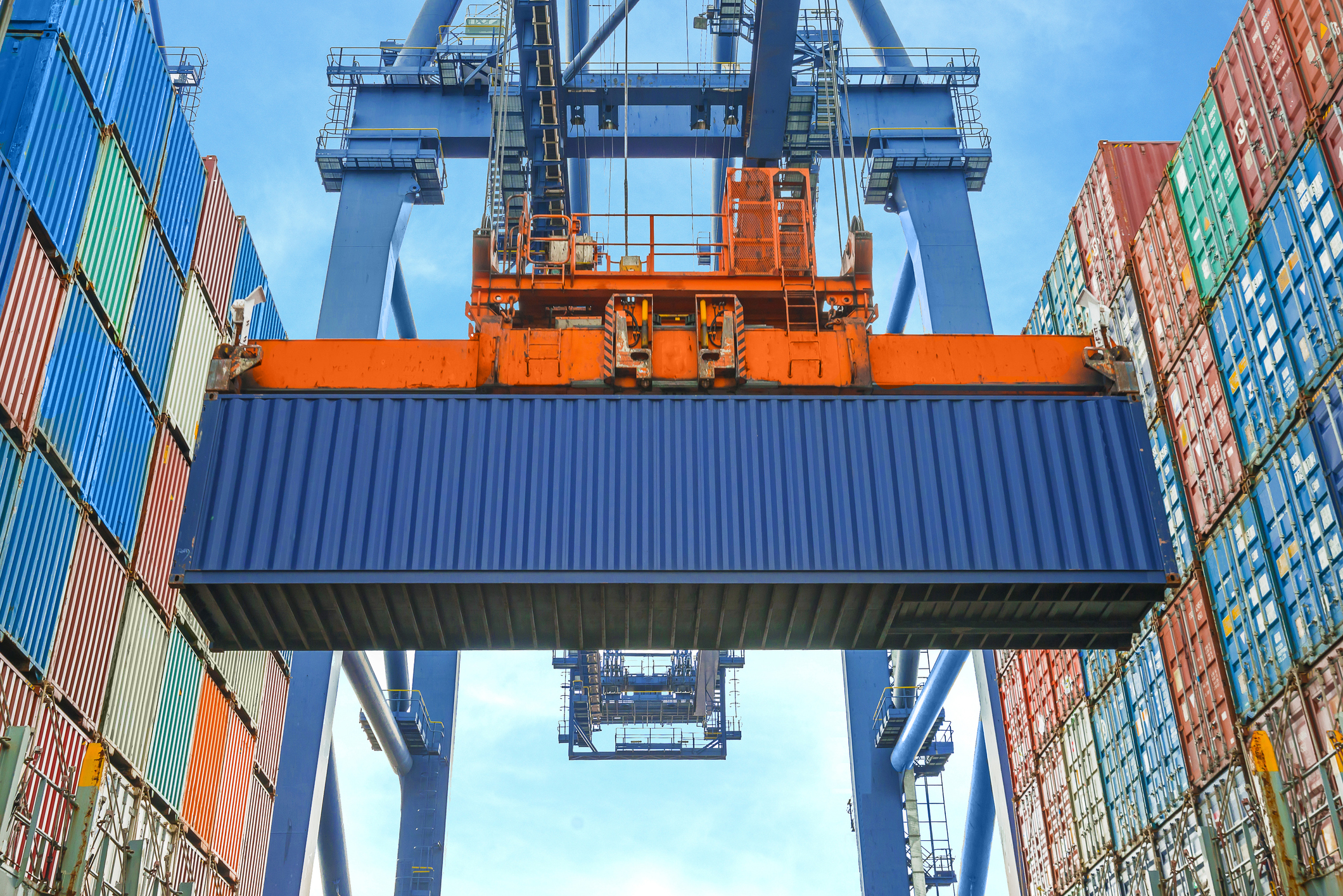 Guindaste portuário movimenta um contêiner azul em um terminal de carga, cercado por pilhas de contêineres coloridos. Estruturas metálicas e céu azul compõem o cenário industrial.