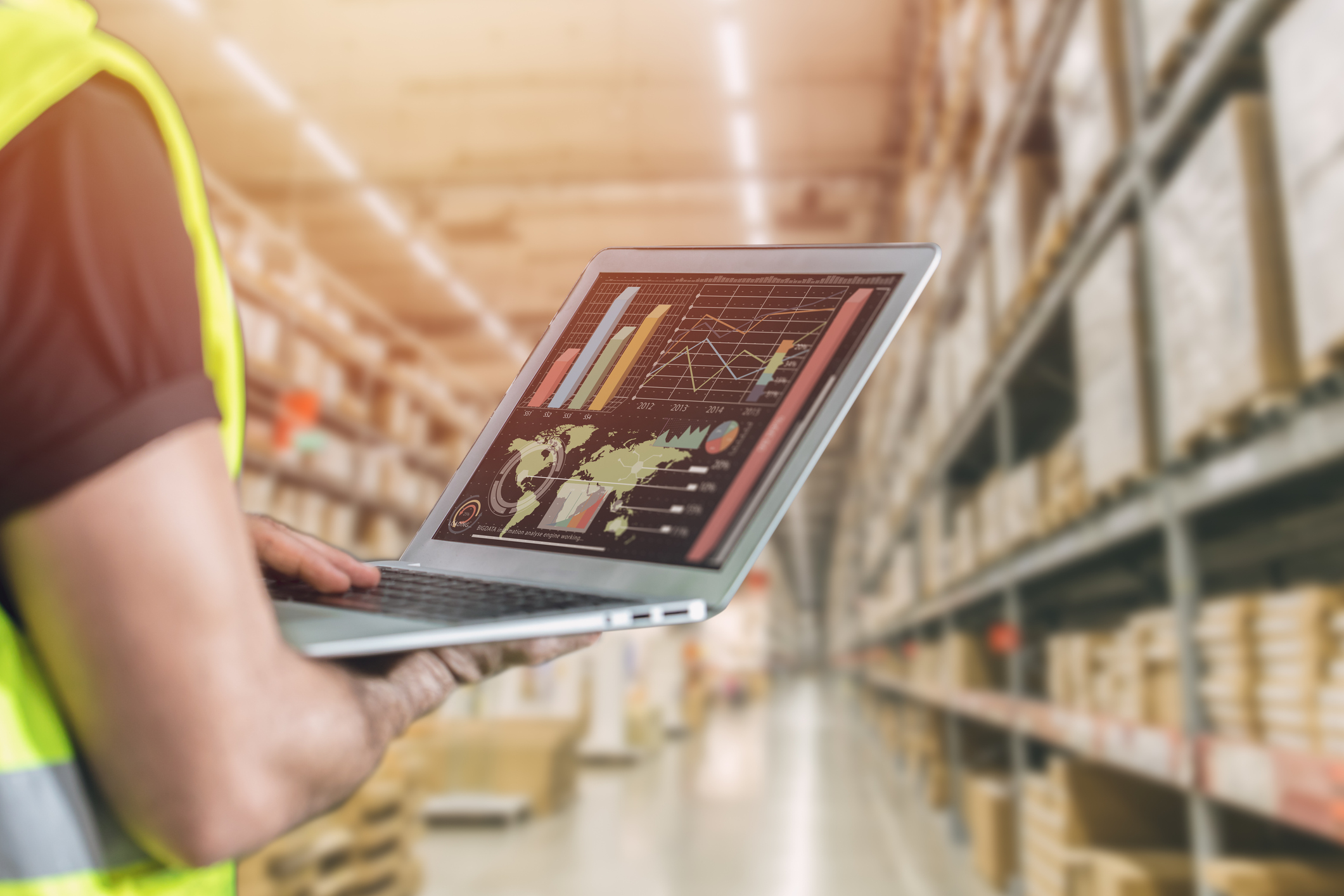 Pessoa com colete de segurança segura um laptop em um armazém. Na tela, gráficos e mapas indicam análise de dados logísticos. Ao fundo, prateleiras organizadas com caixas e iluminação suave.